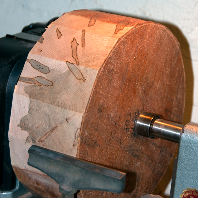 A rough blank of Ambrosia Maple mounted to a wood lathe before turning, workshop visible behind